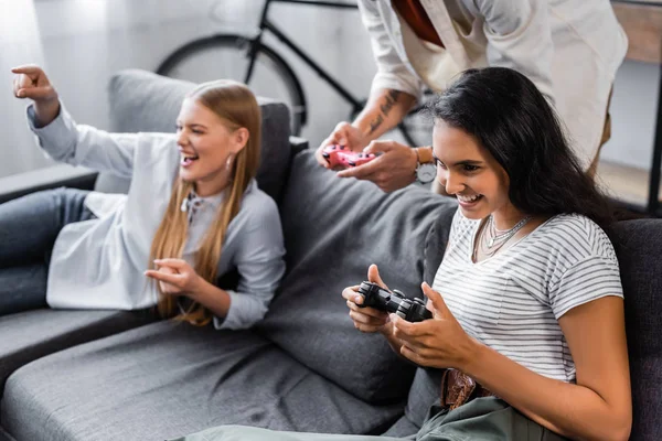 KYIV, UKRAINE - JULY 10, 2019: multicultural friends sitting on sofa and playing video game in apartment — Stock Photo