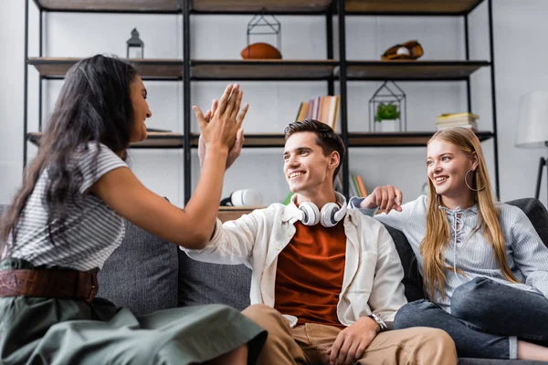 Amigos multiculturales sonriendo y mostrando alta cinco gesto en el apartamento - foto de stock