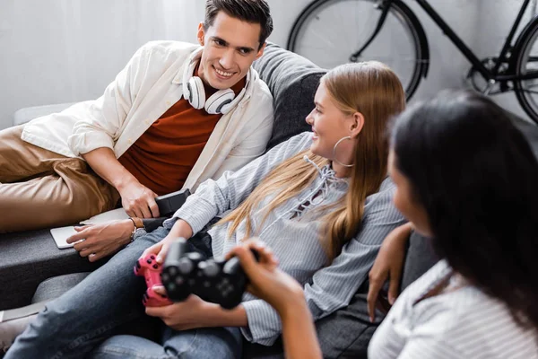 KYIV, UKRAINE - JULY 10, 2019: multicultural friends sitting on sofa and holding joysticks in apartment — Stock Photo