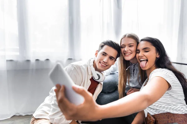 Enfoque selectivo de amigos multiculturales sonriendo y tomando selfie en apartamento - foto de stock