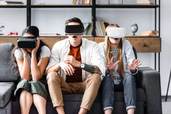 Amigos multiculturales conmocionados con auriculares de realidad virtual sentados en el sofá - foto de stock