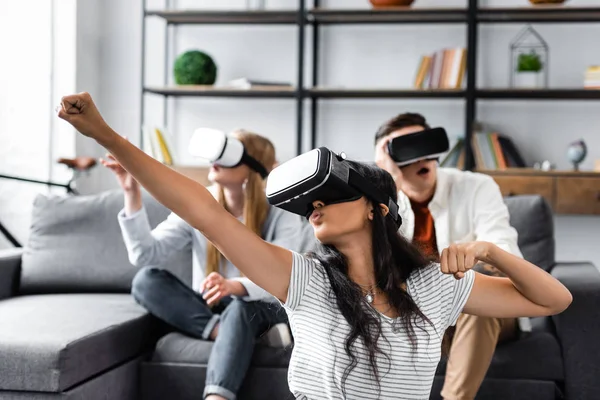 Enfoque selectivo de amigos multiculturales con auriculares de realidad virtual sentados en el sofá - foto de stock