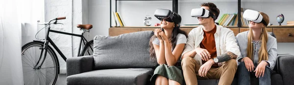 Панорамный снимок мультикультурных друзей с наушниками виртуальной реальности, сидящих на диване — стоковое фото
