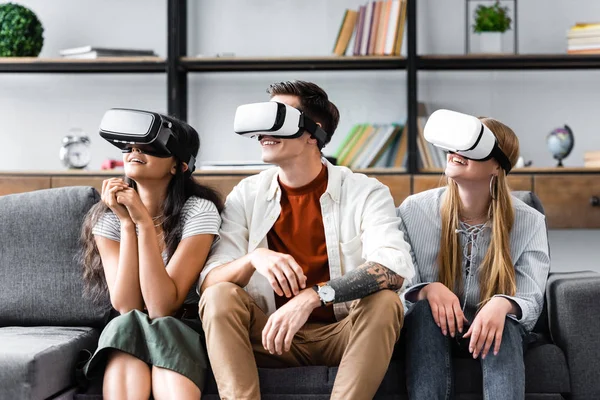 Улыбающиеся мультикультурные друзья с наушниками виртуальной реальности, сидящие на диване — стоковое фото