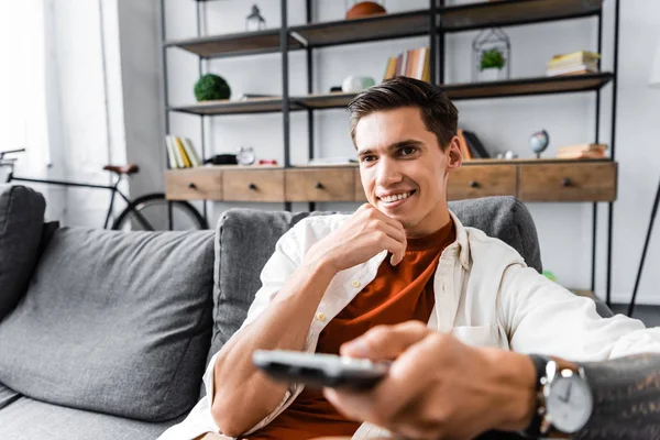 Hombre guapo en camisa sentado en el sofá y sosteniendo el mando a distancia en el apartamento - foto de stock