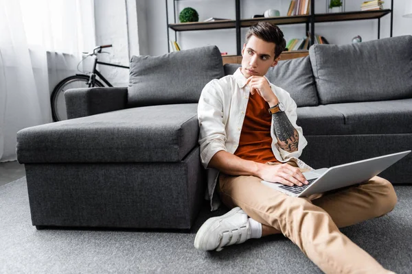 Hombre guapo en camisa sentado en el suelo y el uso de ordenador portátil en el apartamento - foto de stock