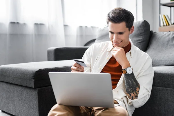 Homem bonito na camisa segurando cartão de crédito e laptop no apartamento — Fotografia de Stock
