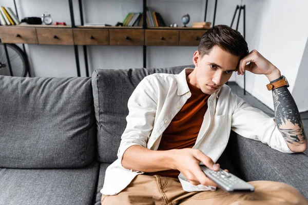 Homem bonito na camisa sentado no sofá e segurando controlador remoto no apartamento — Fotografia de Stock