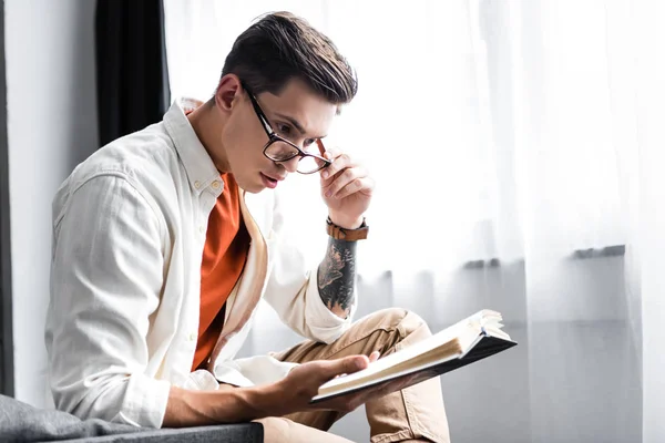 Homem bonito na camisa e óculos livro de leitura no apartamento — Fotografia de Stock
