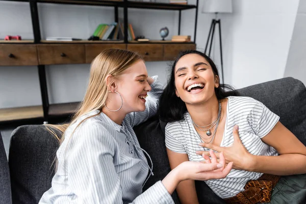 Amigos multiculturales sonriendo y sentados en el sofá en el apartamento - foto de stock