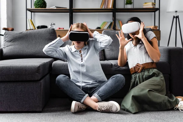 Amigos multiculturales con auriculares de realidad virtual sentados en el piso en el apartamento - foto de stock