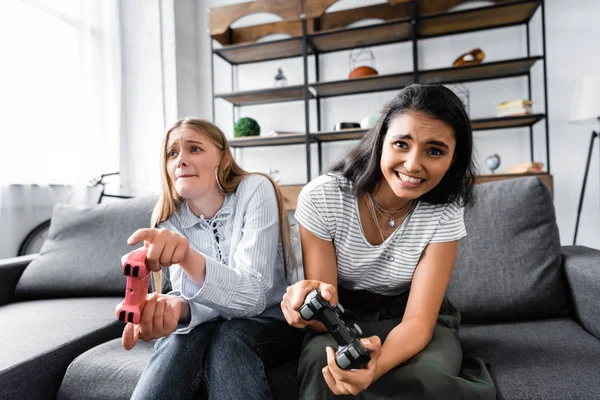 KYIV, UCRANIA - 10 de julio de 2019: amigos multiculturales sentados en el sofá y jugando videojuegos en el apartamento - foto de stock