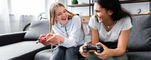 KYIV, UCRANIA - 10 de julio de 2019: plano panorámico de amigos multiculturales sentados en el sofá y jugando videojuegos en el apartamento - foto de stock
