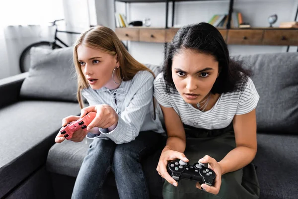 KYIV, UKRAINE - JULY 10, 2019: multicultural friends sitting on sofa and playing video game in apartment — Stock Photo