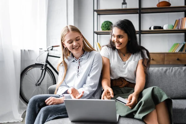 Amigos multiculturais segurando cartão de crédito e usando laptop no apartamento — Fotografia de Stock