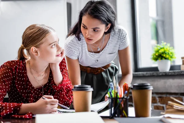 Amigos multiculturales atractivos sentados a la mesa y estudiando en el apartamento - foto de stock