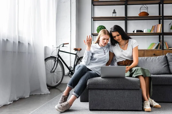 Emotional multicultural friends using laptop and sitting on sofa in apartment — Stock Photo