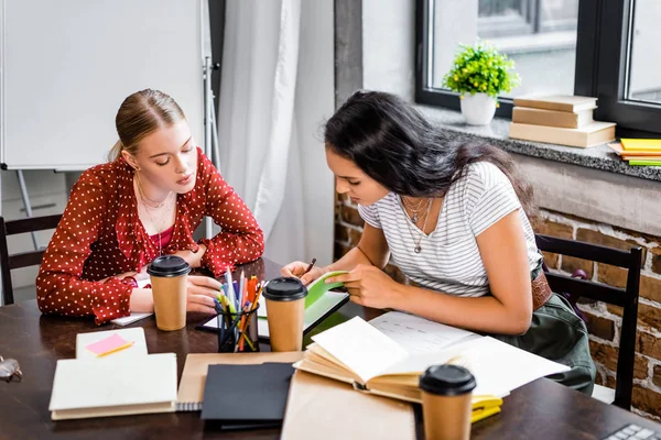Attractive multicultural friends sitting at table and studying in apartment — Stock Photo