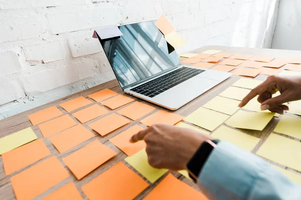 Cropped view of woman pointing with finger at sticky notes near laptop with blank screen on desk — Stock Photo