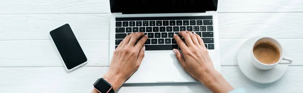 Plano panorámico de la mujer escribiendo en el ordenador portátil cerca de teléfono inteligente y taza de café - foto de stock