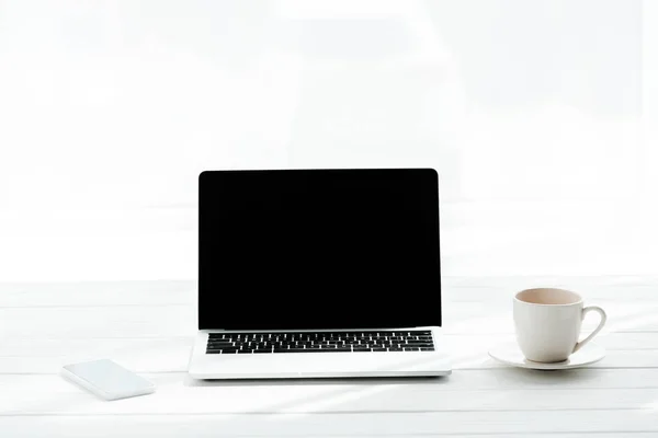 Teléfono inteligente cerca de taza de café y ordenador portátil con pantalla en blanco - foto de stock