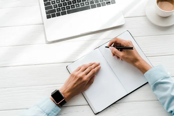 Draufsicht der Frau mit Stift beim Schreiben in Notizbuch in der Nähe von Laptop und Tasse mit Kaffee — Stockfoto