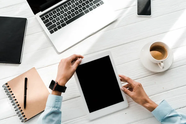 Vista recortada de la mujer sosteniendo tableta digital con pantalla en blanco cerca de gadgets y taza de café - foto de stock