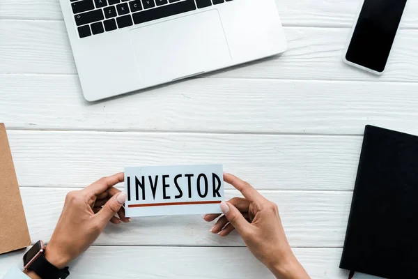 Top view of woman holding card with investor lettering near gadgets and notebooks — Stock Photo