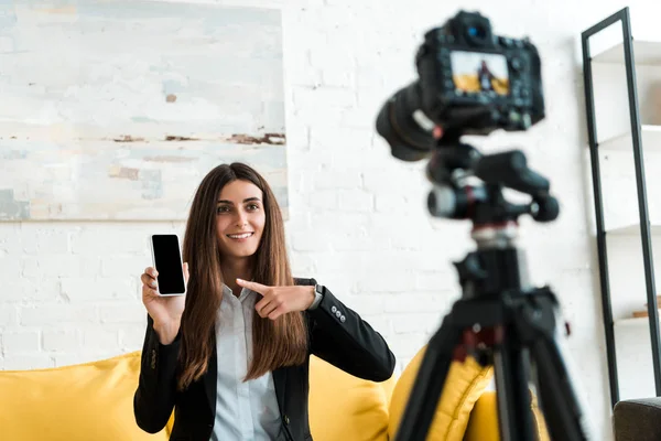 Selektiver Fokus glücklicher Videoblogger, die mit dem Finger auf Smartphone mit leerem Bildschirm in der Nähe der Digitalkamera zeigen — Stockfoto