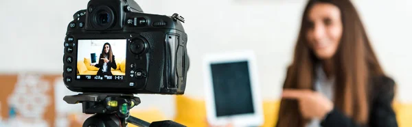 Panoramic shot of digital camera with happy video blogger pointing with finger at digital tablet with blank screen — Stock Photo