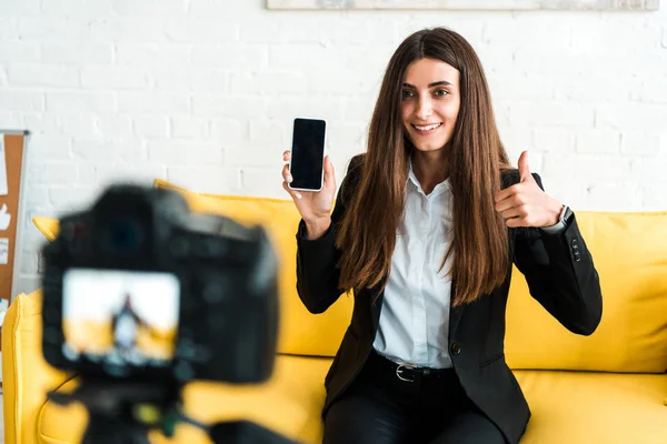 Enfoque selectivo de la mujer feliz mostrando el pulgar hacia arriba y sosteniendo el teléfono inteligente con pantalla en blanco cerca de la cámara digital - foto de stock