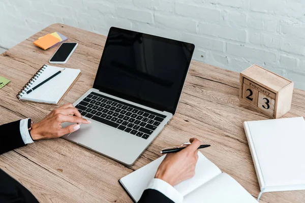 Cropped view of woman holding pen while using laptop with blank screen near smartphone and notebooks — Stock Photo