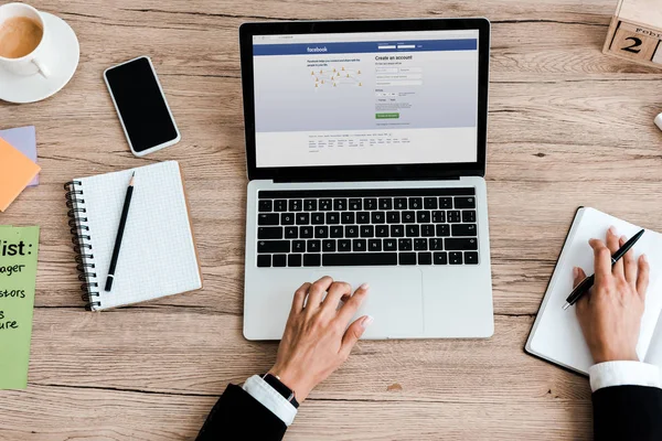 KYIV, UKRAINE - JULY 23, 2019: top view of woman using laptop with facebook website on screen near smartphone with blank screen — Stock Photo