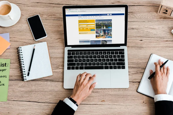 KYIV, UKRAINE - JULY 23, 2019: top view of woman using laptop with booking website near smartphone with blank screen — Stock Photo