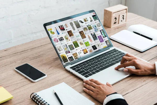 Kyiv, Ukraine - 23. Juli 2019: Ausgeschnittene Ansicht einer Frau, die Laptop mit Pinterest-Website in der Nähe von Smartphone mit leerem Bildschirm und Holzwürfeln benutzt — Stockfoto