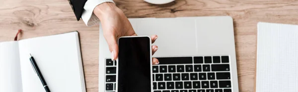 Plano panorámico de la mujer de negocios sosteniendo teléfono inteligente con pantalla en blanco cerca del ordenador portátil - foto de stock