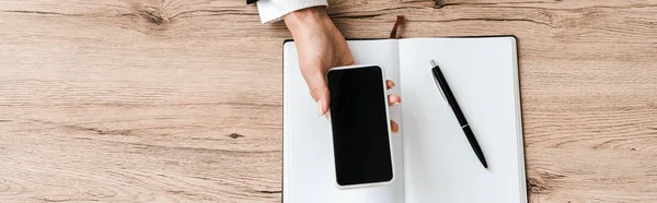 Prise de vue panoramique de femme d'affaires tenant smartphone avec écran vierge près d'un ordinateur portable avec stylo — Photo de stock