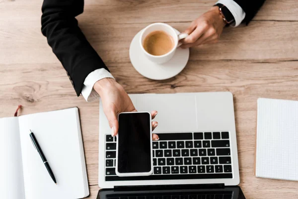 Vista dall'alto della donna d'affari che tiene smartphone con schermo bianco e tazza con caffè vicino al computer portatile — Foto stock