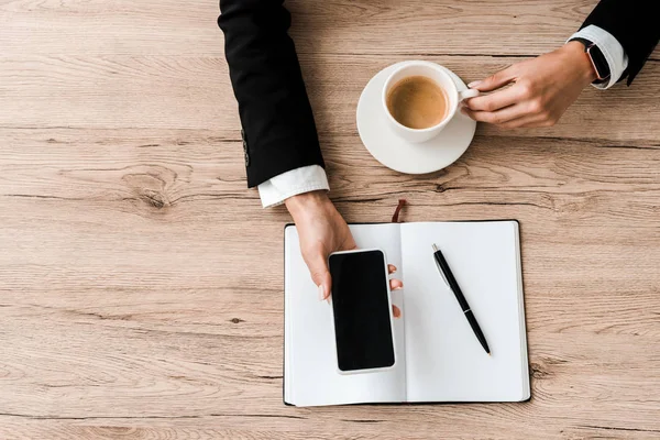 Vista dall'alto della donna d'affari che tiene smartphone con schermo bianco e tazza con caffè vicino al notebook con penna — Foto stock