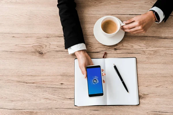 KYIV, UKRAINE - JULY 23, 2019: top view of businesswoman holding smartphone with shazam app on screen and cup with coffee near notebook and pen — Stock Photo