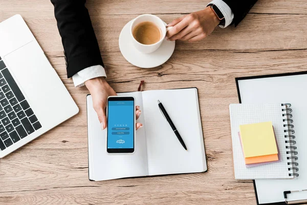 KYIV, UCRANIA - 23 de julio de 2019: vista superior de la mujer de negocios que sostiene el teléfono inteligente con la aplicación skype en la pantalla y la taza con café cerca del portátil y la pluma - foto de stock