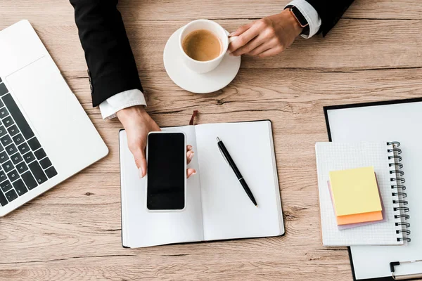 Vista dall'alto della donna d'affari che tiene smartphone con schermo bianco e tazza con caffè vicino a notebook e penna — Foto stock