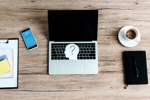 KYIV, UKRAINE - JULY 23, 2019: top view of smartphone with skype app near laptop with blank screen and paper human head with question mark — Stock Photo