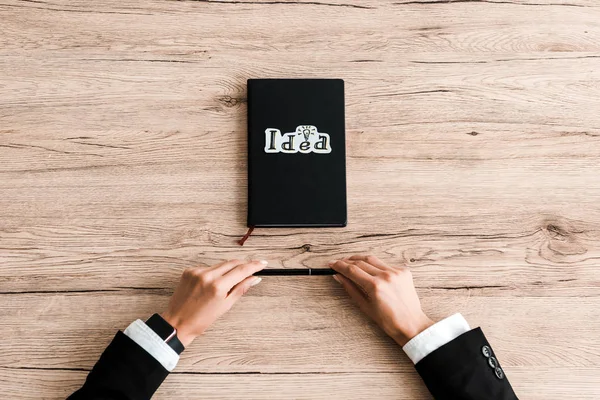 Cropped view of woman holding pen near paper with idea lettering on black notebook — Stock Photo