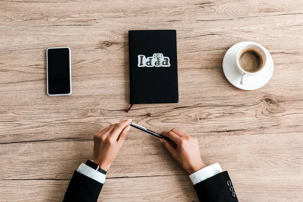 Ausgeschnittene Ansicht einer Frau mit Stift in Papiernähe mit Ideenschrift auf schwarzem Notizbuch und Tasse Kaffee — Stockfoto