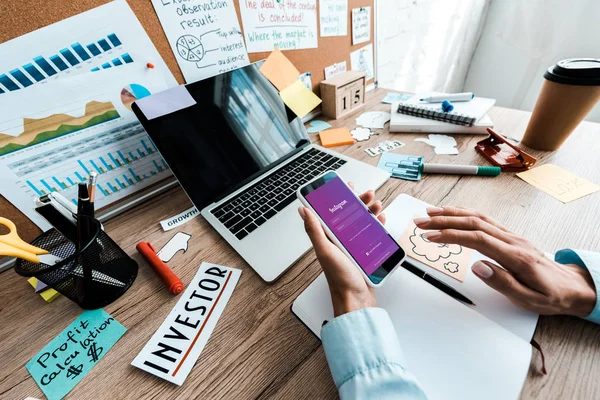 KYIV, UCRANIA - 23 de julio de 2019: vista recortada de una mujer de negocios sosteniendo un teléfono inteligente con aplicación instagram cerca de un portátil y una computadora portátil con pantalla en blanco - foto de stock