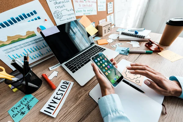 KYIV, UKRAINE - JULY 23, 2019: cropped view of businesswoman holding iphone near notebook and laptop with blank screen — Stock Photo