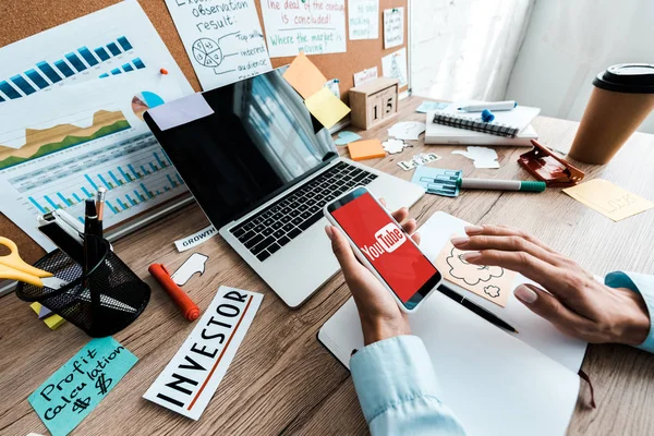 KYIV, UKRAINE - JULHO 23, 2019: visão recortada de empresária segurando smartphone com aplicativo youtube perto de notebook e laptop com tela em branco — Fotografia de Stock