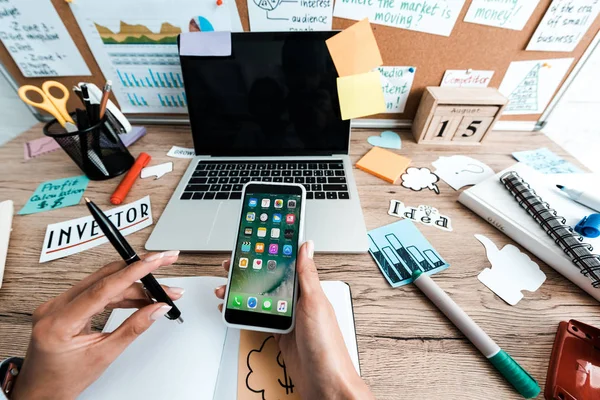 KYIV, UKRAINE - JULY 23, 2019: cropped view of businesswoman holding iphone near notebook, sticky notes and laptop with blank screen — Stock Photo