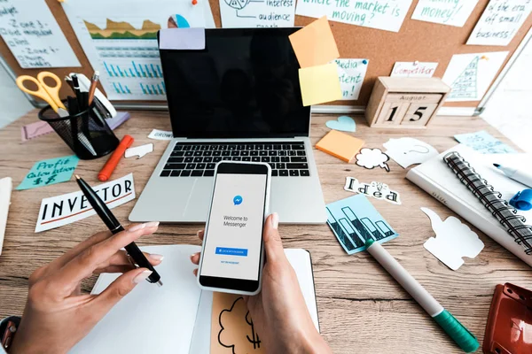 KYIV, UCRANIA - 23 de julio de 2019: vista recortada de una mujer sosteniendo un teléfono inteligente con aplicación de mensajería cerca de notas adhesivas con letras y computadora portátil con pantalla en blanco - foto de stock